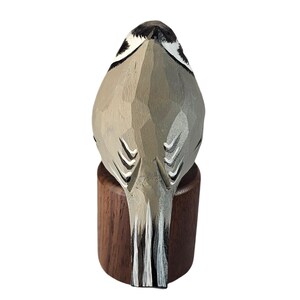 May include: A carved wooden bird sculpture, viewed from the back, perched on a small, dark wooden base. The bird is primarily gray with black and white accents, showcasing detailed carving and a natural aesthetic.