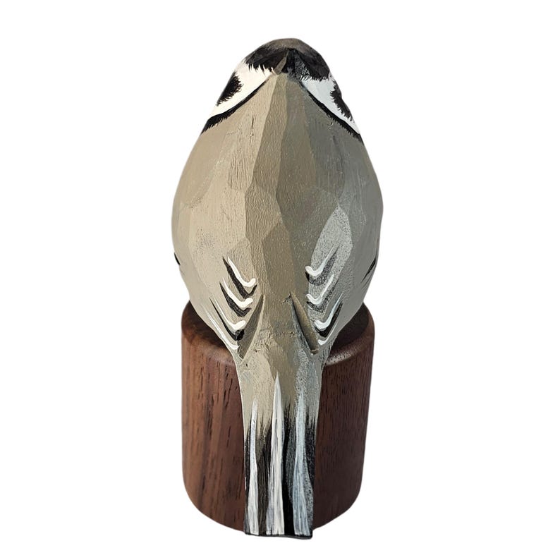 May include: A carved wooden bird sculpture, viewed from the back, perched on a small, dark wooden base. The bird is primarily gray with black and white accents, showcasing detailed carving and a natural aesthetic.