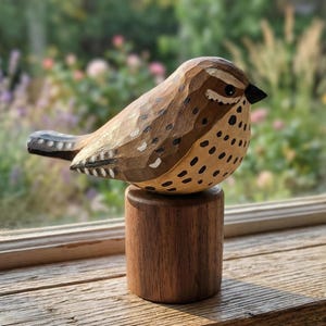 以下が含まれることがあります： 茶色、ベージュ、黒の色合いの手彫りの木製鳥の彫刻。小さな木製の台座に置かれています。鳥は、白い点や黒いくちばしなど、詳細な彫刻と塗装されたアクセントが特徴です。彫刻は木製の表面に展示されています。