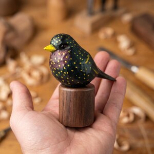 May include: A hand-carved wooden bird figurine. The bird has a dark green body with yellow spots, a yellow beak, and a purple neck. It is mounted on a small, cylindrical wooden base. The bird is approximately 7.6 cm long.