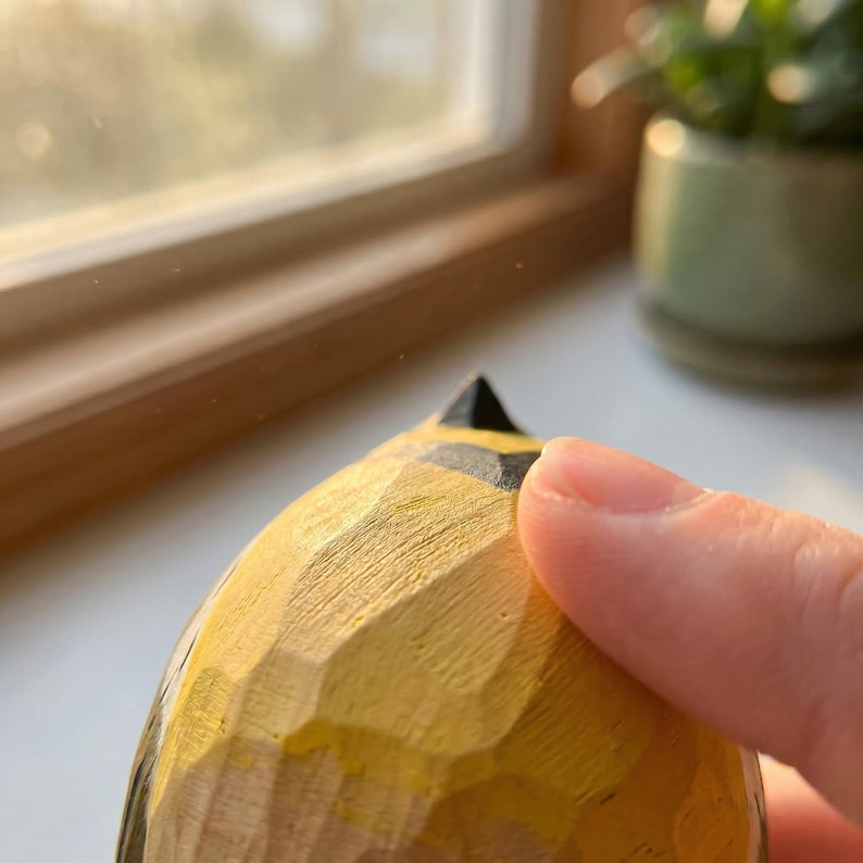 Close-up of a hand-carved wooden Western Meadowlark figurine held between two fingers to show its miniature scale. The 3.5-inch bird features a faceted whittled texture and hand-painted yellow and black details, making it a perfect rustic desk accessory, nature-themed shelf sitter, or unique gift for bird watchers.