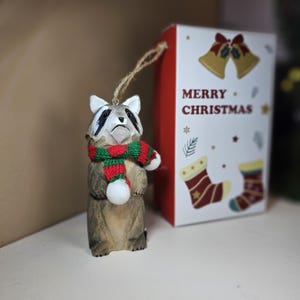 Op de afbeelding: Houten wasbeer ornament met een rood-groene sjaal. Het ornament hangt aan een touwtje en heeft een witte pompon aan het uiteinde. Het ornament bevindt zich voor een rood-witte doos met de tekst "Merry Christmas".