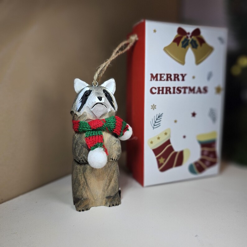 Op de afbeelding: Houten wasbeer ornament met een rood-groene sjaal. Het ornament hangt aan een touwtje en heeft een witte pompon aan het uiteinde. Het ornament bevindt zich voor een rood-witte doos met de tekst "Merry Christmas".