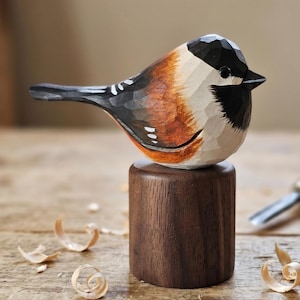 Puede incluir: Figurita de pájaro de madera tallada a mano con cabeza negra, cuerpo blanco y naranja y cola negra, posada sobre un pequeño soporte de madera oscura. La escultura de pájaro está detallada con la veta de la madera visible y se asienta sobre una base cilíndrica.