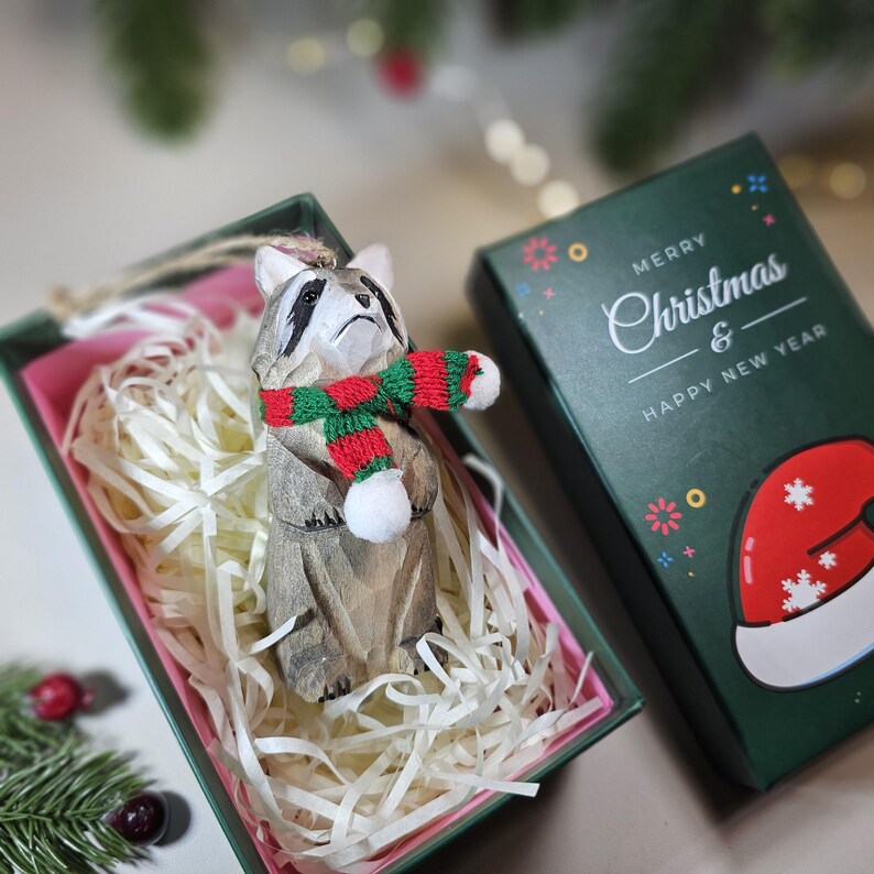 Hand-carved wooden raccoon ornament wearing a red and green knit scarf inside a green holiday gift box. Basswood woodland animal tree decor.