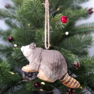 K&ouml;nnte beinhalten: Ein h&ouml;lzerner Waschb&auml;r-Anh&auml;nger mit einem grau-braun gestreiften Schwanz, der an einer Bindfadenkordel an einem Weihnachtsbaum h&auml;ngt.