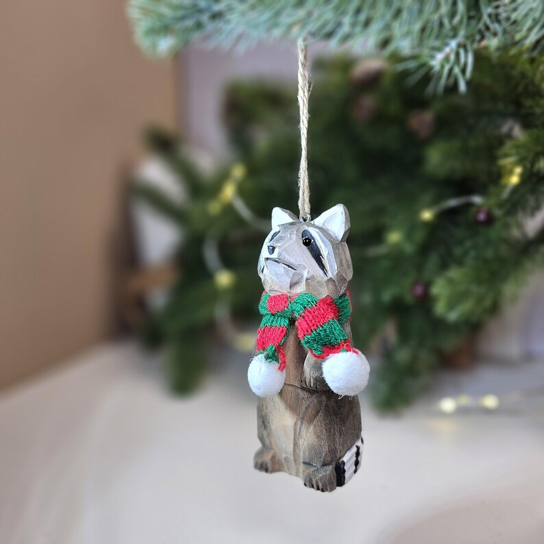 Op de afbeelding: Houten kerstversiering in de vorm van een wasbeer met een rood-groene gestreepte sjaal en witte pompoenen.
