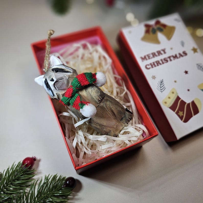 Hand-carved wooden raccoon ornament wearing a red and green knit scarf inside a "Merry Christmas" gift box. Basswood woodland animal tree decor.