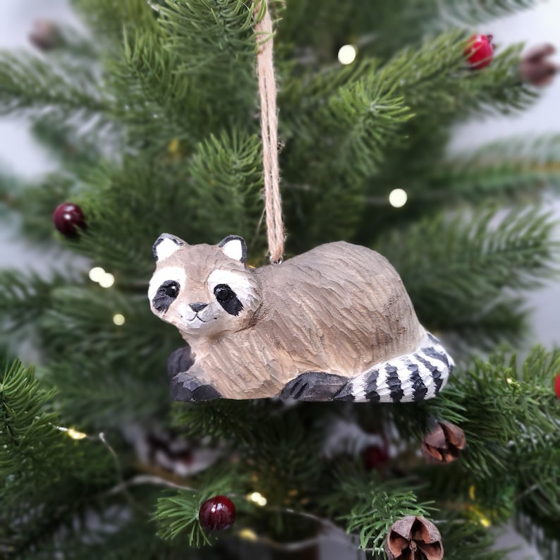 Hand-carved wooden raccoon ornament hanging on a Christmas tree. Hand-painted basswood wildlife animal figurine for holiday woodland decor.