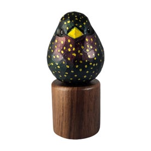 May include: A wooden bird ornament, hand-carved and painted. The bird has a dark body with yellow spots, a yellow beak, and a purple hue. It is mounted on a cylindrical wooden base. The base is made of dark wood.