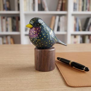 Side profile view of a hand-carved wooden European Starling figurine showcasing its intricate, hand-painted yellow speckles over glossy dark iridescent green and purple plumage. The realistic basswood bird sculpture features a bright yellow beak and is perched on a round black walnut base, displayed on a wooden desk next to a black and gold fountain pen. Perfect for nature-inspired home decor, office desk styling, or a unique gift for bird watchers.