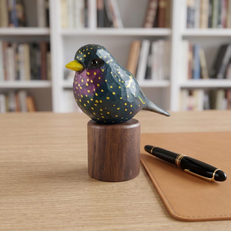 Side profile view of a hand-carved wooden European Starling figurine showcasing its intricate, hand-painted yellow speckles over glossy dark iridescent green and purple plumage. The realistic basswood bird sculpture features a bright yellow beak and is perched on a round black walnut base, displayed on a wooden desk next to a black and gold fountain pen. Perfect for nature-inspired home decor, office desk styling, or a unique gift for bird watchers.