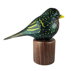May include: A hand-carved wooden bird figurine. The dark green body is speckled with yellow dots, with yellow wing markings and a yellow beak. It is mounted on a small, round, dark brown wooden base.