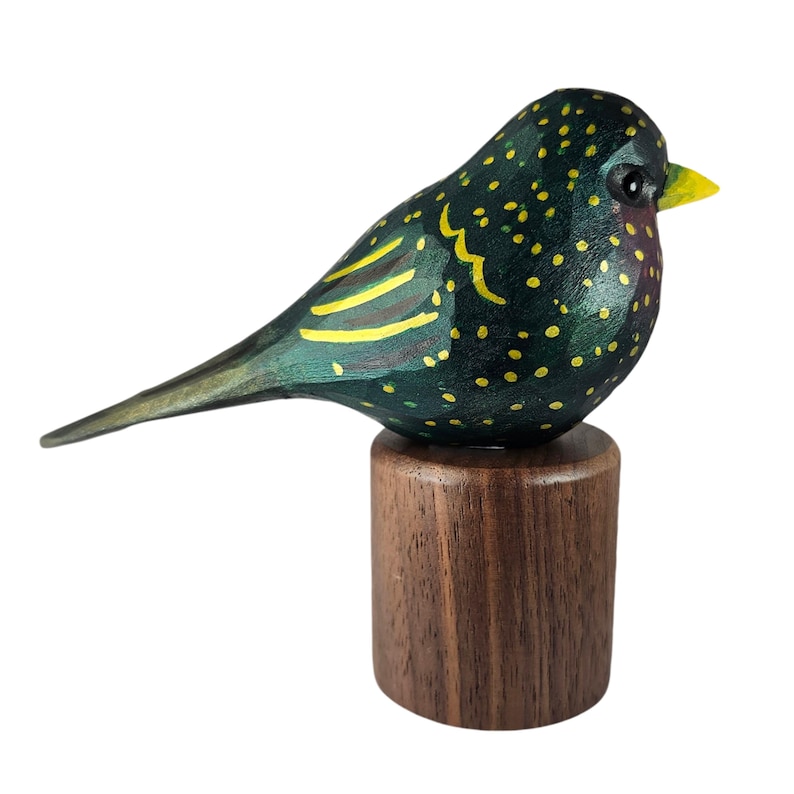 May include: A hand-carved wooden bird figurine. The dark green body is speckled with yellow dots, with yellow wing markings and a yellow beak. It is mounted on a small, round, dark brown wooden base.