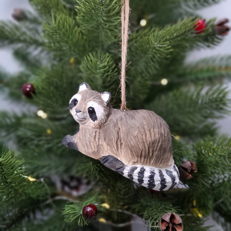 K&ouml;nnte beinhalten: Ein h&ouml;lzerner Waschb&auml;r-Anh&auml;nger mit braunem und grauem K&ouml;rper und einem schwarz-wei&szlig; gestreiften Schwanz. Der Anh&auml;nger h&auml;ngt an einer braunen Schnur.