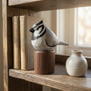 Hand-painted Bridled Titmouse wood figurine on a shelf by vintage books and a vase. Basswood bird sculpture for rustic home decor.