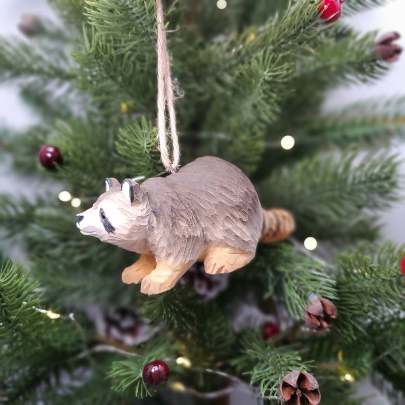 K&ouml;nnte beinhalten: Ein h&ouml;lzerner Waschb&auml;r-Anh&auml;nger, der an einem Weihnachtsbaum h&auml;ngt. Der Waschb&auml;r ist braun und grau mit einem langen Schwanz und einer schwarzen Nase.