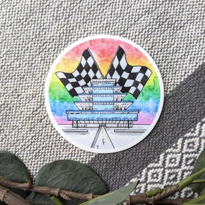 May include: A watercolor illustration of the Indianapolis Motor Speedway, featuring a rainbow background and checkered flags. The iconic track is depicted in detail, with the start/finish line and the iconic pagoda.