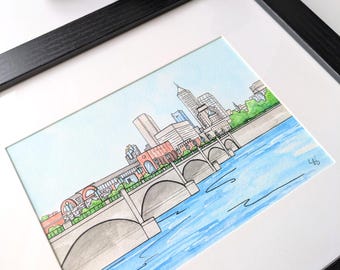 Puede incluir: Una pintura de acuarela de una ciudad con un puente sobre un río. Los edificios están en el fondo y el puente está en primer plano. La pintura está enmarcada en un marco negro.