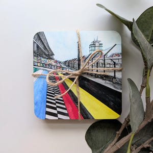 May include: A set of square coasters tied with twine. Each coaster features a watercolor illustration of a racetrack with a blue sky background. The track has red, yellow, and black stripes, and a checkered pattern.
