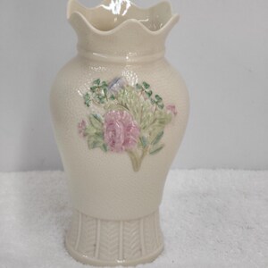 May include: Cream-colored ceramic vase with a textured surface and a ruffled top. The vase features a floral design with pink roses and green leaves. The base has a textured, basket-weave design.