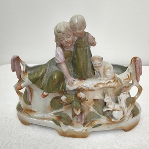 Puede incluir: Un centro de mesa de porcelana con dos niños vestidos de verde oliva sentados sobre una base decorativa con un gato y una ardilla. La base está adornada con hojas y ramas esculpidas, acabada en tonos apagados de verde, marrón y rosa suave.