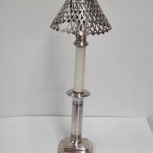 Peut inclure: Un bougeoir argenté avec un abat-jour ajouré décoratif. Le bougeoir a une base carrée, une longue tige cylindrique et un porte-bougie circulaire. L'abat-jour est en forme de cône et présente un motif géométrique détaillé.