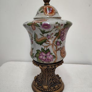 Puede incluir: Un tarro de cerámica con tapa sobre una base blanca, decorado con un colorido patrón floral de flores rosas, naranjas y verdes sobre ramas. El tarro se asienta sobre una base de pedestal color bronce oscuro con tallas de hojas y una tapa a juego.