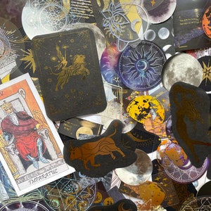 May include: A collection of celestial-themed stickers and tarot cards. The stickers feature gold foil accents with moons, stars, and zodiac symbols. One tarot card displays a king, while another shows a winged figure riding a lion.