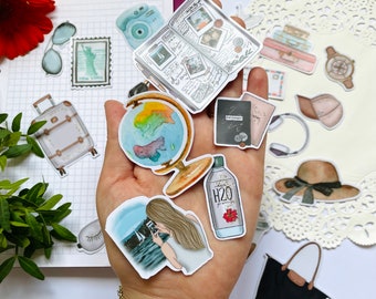 Op de afbeelding: Een verzameling stickers met een reisthema, waaronder een wereldbol, paspoort, camera, koffer en dagboek. De stickers zijn in verschillende kleuren, met een aquarel-stijl. De afbeelding bevat ook een zwarte draagtas en een zonnehoed.