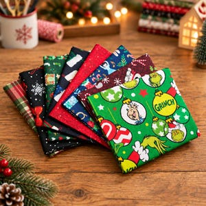 May include: A stack of folded fabric squares featuring various Christmas-themed patterns, including green fabric with Grinch illustrations and ornaments, red and black plaid, dark blue with snowflakes, and green with white holiday motifs.