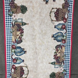 May include: Rectangular kitchen towel with a cream-colored center and a border featuring a blue and white checkered pattern. The towel is decorated with images of fruit baskets, bottles, and grapes, with a red trim.