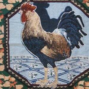May include: A tapestry featuring a colorful rooster with a blue, brown, and white patterned tail. The rooster is standing on a blue and white patterned background with white flowers. The tapestry is framed with a brown and green border.