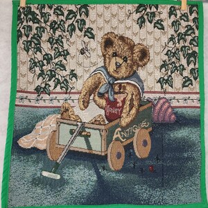 May include: A brown teddy bear wearing a blue sailor shirt and holding a red jar labeled "Honey" sits in a wooden wagon with green wheels. The wagon is on a green and blue background with ivy vines and a white and brown patterned wall.
