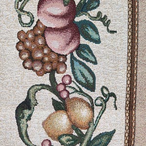 May include: Close-up of a tapestry with a repeating pattern of fruits and foliage. The design features peaches, grapes, lemons, and berries in shades of pink, brown, yellow, and green against a cream background. The border has a brown geometric pattern.
