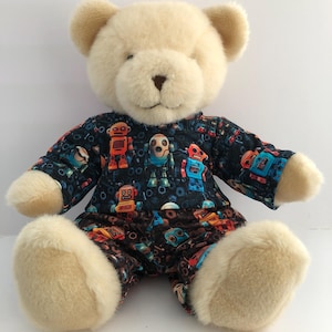 Può includere: Un orsacchiotto di peluche color crema, con un pigiama blu scuro decorato con illustrazioni di robot colorati. L'orso ha un naso marrone ed è seduto.
