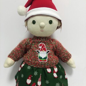 May include: A handmade doll dressed for Christmas. The doll wears a red and white Santa hat with a white pom-pom, a red and green sweater with a gnome applique, and a green skirt with gnome and snowflake patterns. The doll has green eyes.