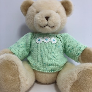 Puede incluir: Un oso de peluche marrón claro con un suéter de punto verde con flores blancas y centros azules. El suéter tiene un patrón moteado.