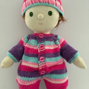 Könnte beinhalten: Eine handgefertigte Puppe mit einem gestrickten Outfit und Hut. Das Outfit ist in Rosa-, Lila- und Türkistönen gestreift und hat passende Knöpfe. Der Hut ist ebenfalls in passenden Farben gestreift und hat einen kleinen weißen Bommel. Die Puppe hat grüne Augen und braune Haare.