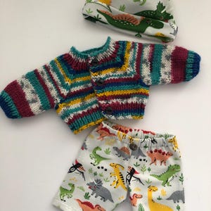 May include: A baby clothing set featuring a knitted, multicolored striped sweater with button closures, matching shorts with a dinosaur print, and a dinosaur-themed hat. The set is designed for infants.