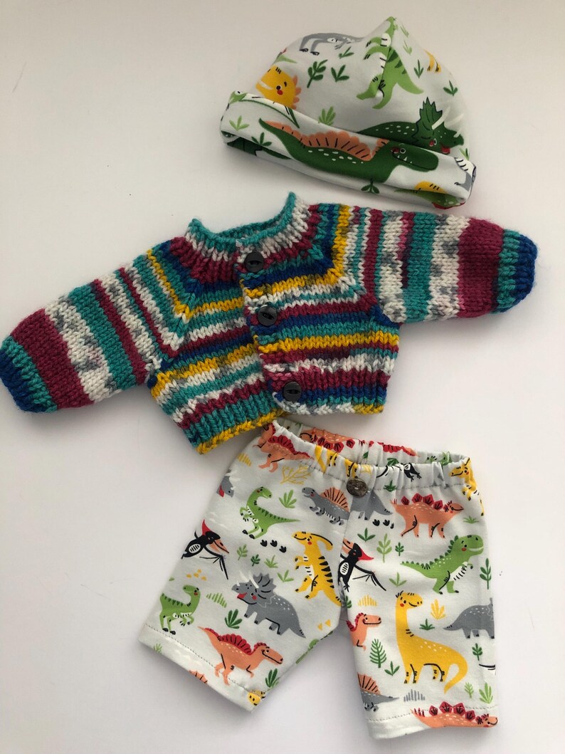 May include: A baby clothing set featuring a knitted, multicolored striped sweater with button closures, matching shorts with a dinosaur print, and a dinosaur-themed hat. The set is designed for infants.