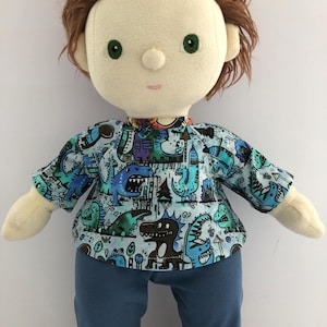 Op de afbeelding: Handgemaakte pop met een lichte huidskleur, groene ogen en bruin haar. De pop draagt een blauw shirt met een dinosauruspatroon en een blauwe broek. De schoenen van de pop zijn bruin.