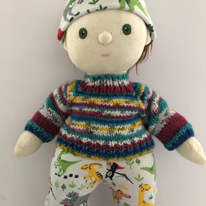 May include: A handmade doll wearing a colorful striped sweater, dinosaur-print pants, and a matching hat. The doll has green eyes and brown hair. The doll is approximately 12 inches tall.