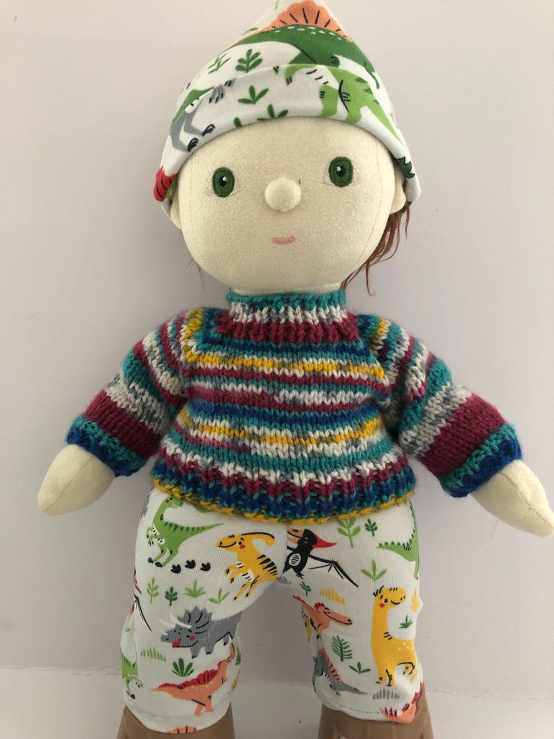 May include: A handmade doll wearing a colorful striped sweater, dinosaur-print pants, and a matching hat. The doll has green eyes and brown hair. The doll is approximately 12 inches tall.