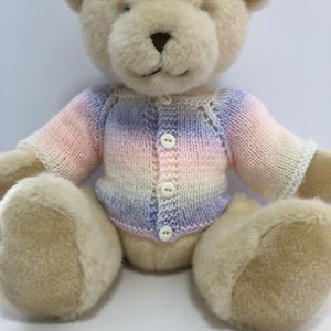 May include: A white teddy bear wearing a knitted cardigan with a pink, white and purple ombre pattern. The cardigan has a button fastening and a delicate lace pattern around the neckline.