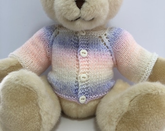 May include: A white teddy bear wearing a knitted cardigan with a pink, white, and purple ombre pattern. The cardigan has a button closure and a delicate lace pattern around the neckline.