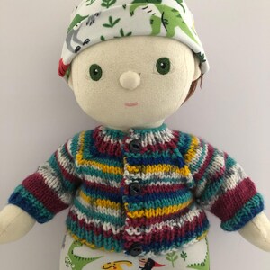 May include: A handmade doll wearing a knitted, striped cardigan in shades of blue, yellow, and burgundy. The doll has a matching hat with a dinosaur print and pants with a similar dinosaur pattern. The doll has green eyes and brown hair.