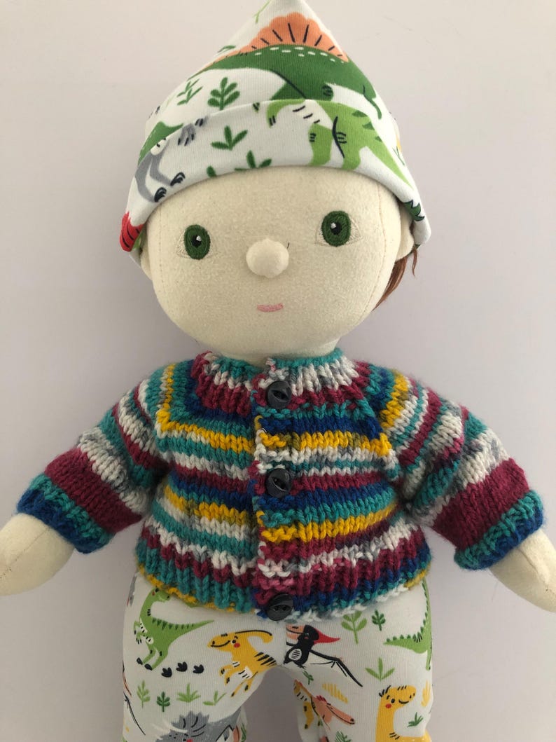 May include: A handmade doll wearing a knitted, striped cardigan in shades of blue, yellow, and burgundy. The doll has a matching hat with a dinosaur print and pants with a similar dinosaur pattern. The doll has green eyes and brown hair.
