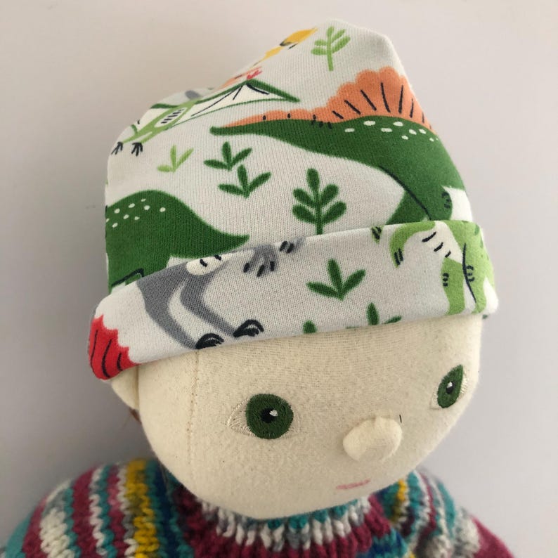 May include: A white beanie hat with a green dinosaur pattern, worn by a doll. The hat features a folded brim and a colorful knit sweater. The dinosaur design includes green dinosaurs, gray animals, and green foliage.