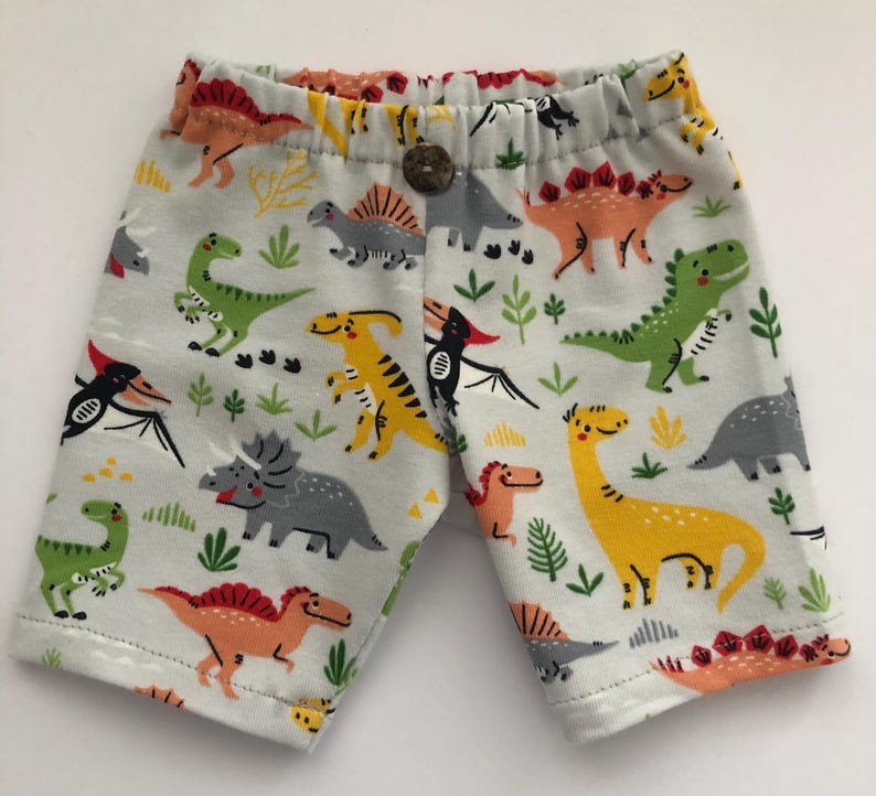 May include: Light gray shorts with an all-over dinosaur print in various colors. The dinosaurs are orange, yellow, green, and gray. The shorts have an elastic waistband with a wooden button closure.